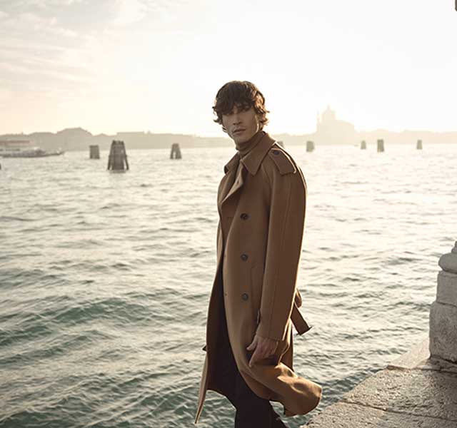 Bugatti Camel Overcoat – Winter 2025 Collectie Man in een stijlvolle Bugatti zomeroutfit met lichte broek en reisbag, staand in een zonnige villa-ingang.