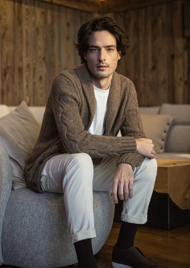 Man draagt de Gran Sasso Cable Cardigan 10189/19107-130, verkrijgbaar bij Van den Broek Herenmode in Haarlem, zittend in warme interieursetting.